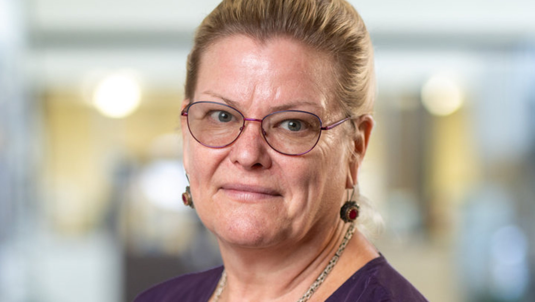 Frau mit kurz geschnittenen, hellblonden Haaren und Brille lächelt freundlich. Sie trägt ein lila langärmliges Oberteil und eine Halskette mit Ohrringen. Im Hintergrund ein helles, modernes Büro mit Regalen und Türen.