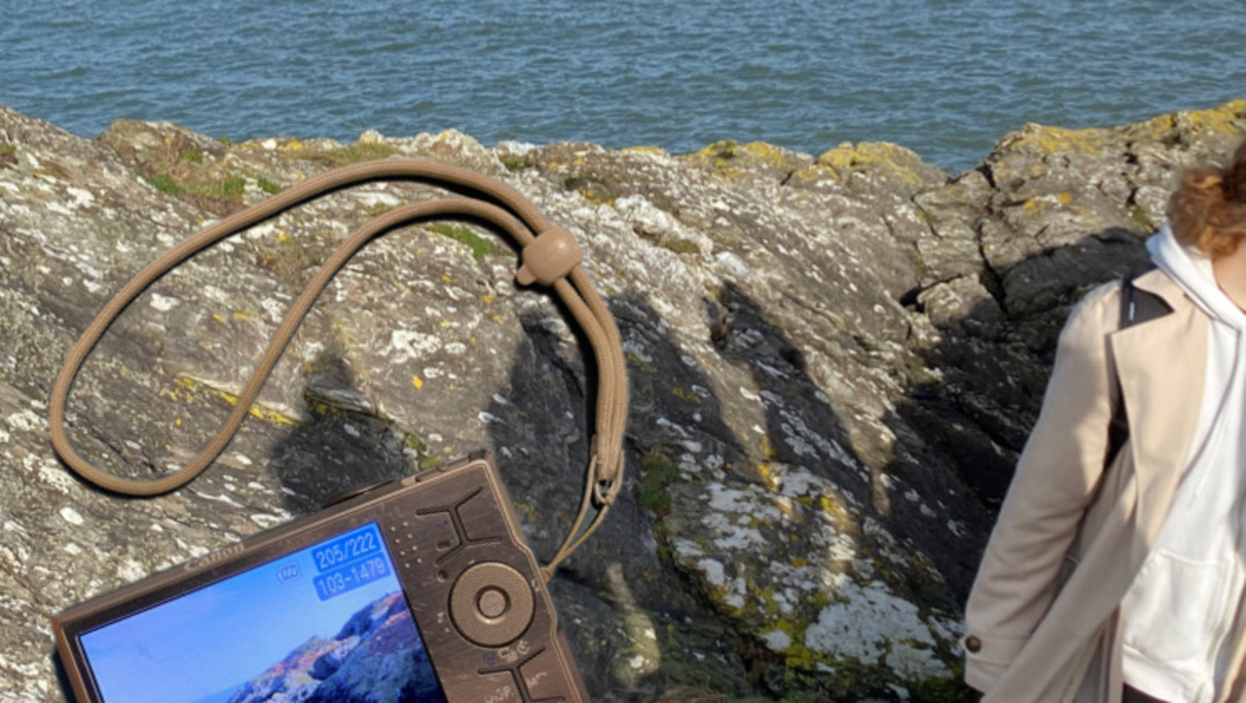 Person hält ein altes Handy mit Landschaftsfotos vor einer steinernen Aussichtsplattform auf einer Klippe. Im Hintergrund ist das Meer zu sehen. Das Gerät zeigt zwei Bilder: eins mit einer Burgruine und eins mit einer grünen Hügellandschaft.