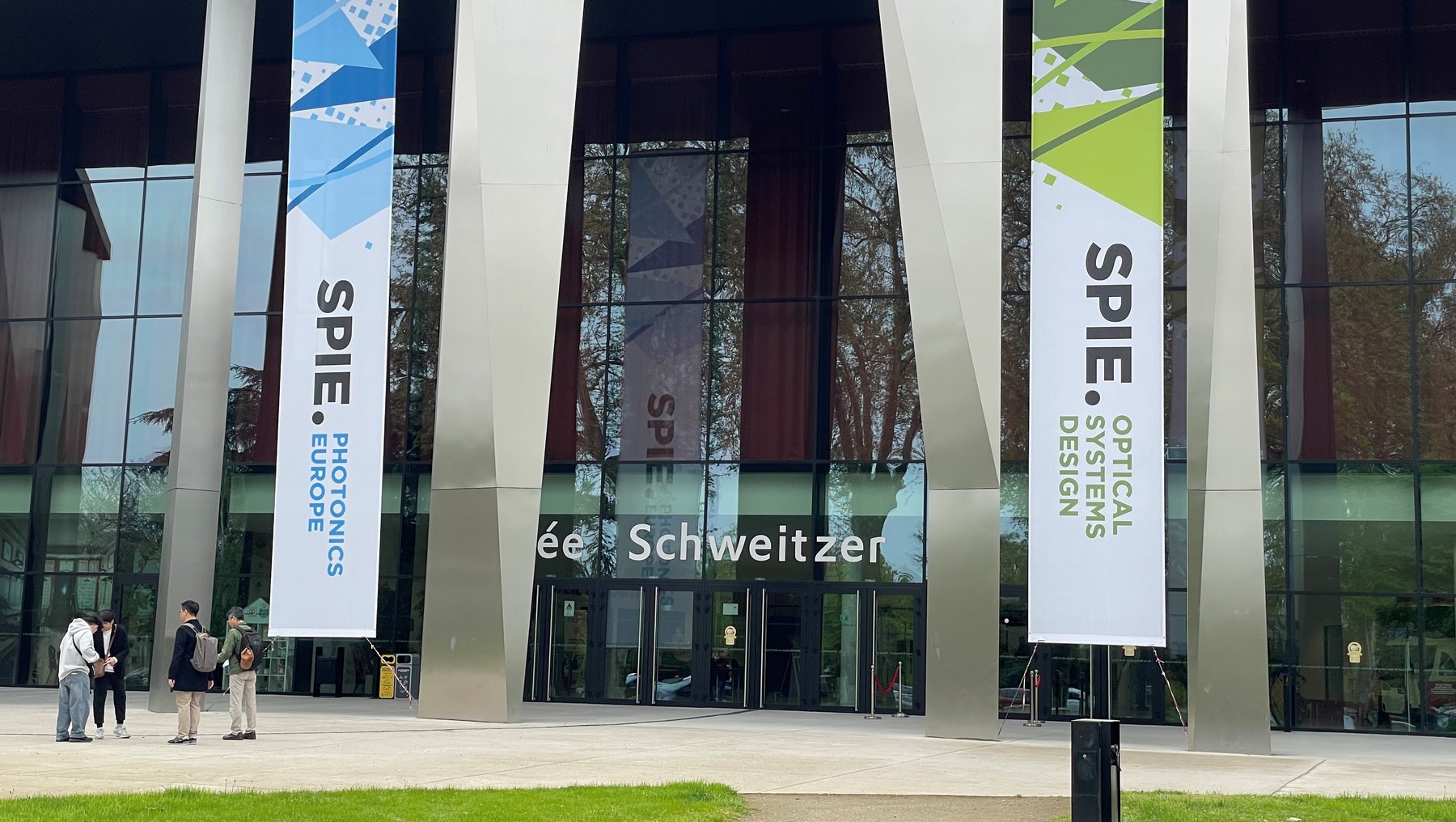 Moderne Glasfassade eines Gebäudes mit drei großen Bannerwerbungen für SPIE, die Themen Optische Systeme, Elektronik und Systemdesign behandeln. Vor dem Eingang stehen zwei Personen und betreten das Gebäude.