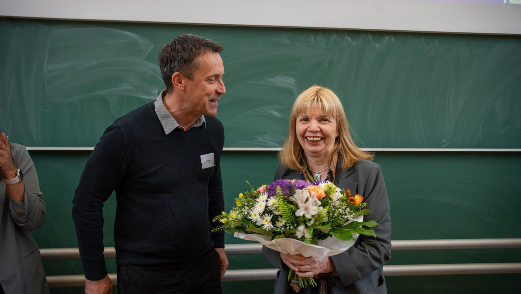 Eine lächelnde Frau hält einen Strauß mit bunten Blumen und Grünpflanzen. Neben ihr steht ein Mann, beide wirken zufrieden und scheinen eine feierliche oder erfolgreiche Situation zu teilen.