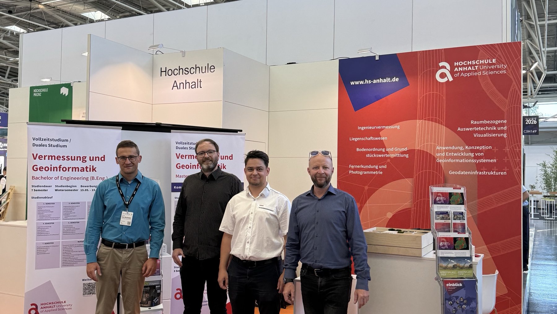 Vier Personen stehen lächelnd auf einer Messe vor einem Stand der Hochschule Anhalt. Der Stand thematisiert Vermessung und Geoinformatik. Im Hintergrund sind Informationsmaterialien und ein roter Bereich mit Logo und Text zu sehen.