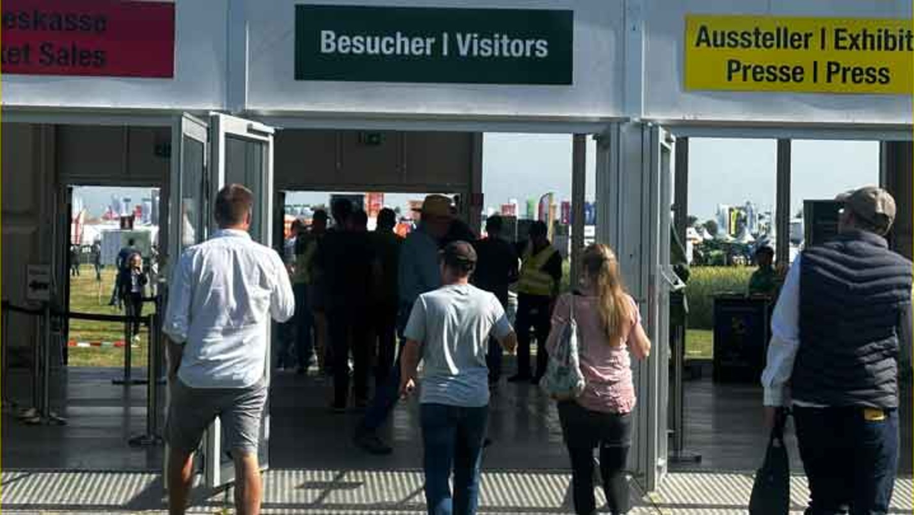 Menschen betreten eine Messe durch zwei getrennte Eingänge: links für Besucher und rechts für Aussteller sowie Presse.
