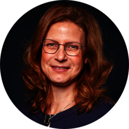 Porträt einer lächelnden Person mit hellbraunen, welligen Haaren, die eine Brille trägt und ein dunkles Oberteil mit einem silbernen Anhänger um den Hals.