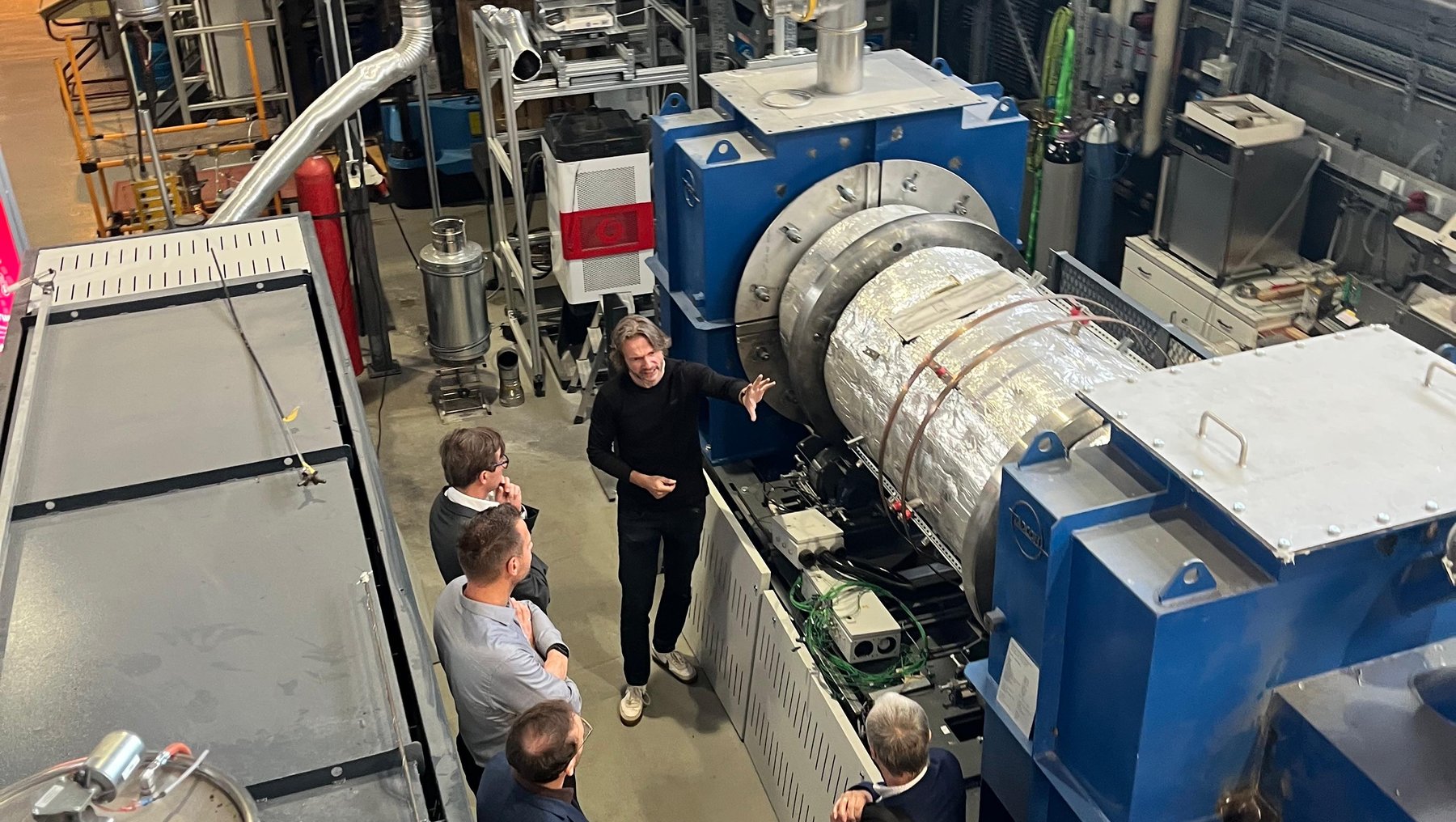 Gruppe von Personen in einer Industriehalle, die eine große technische Anlage mit Rohren, Ventilen und einem horizontalen zylindrischen Behälter besichtigen. Ein Mitarbeiter erklärt die Funktionsweise der Maschine.