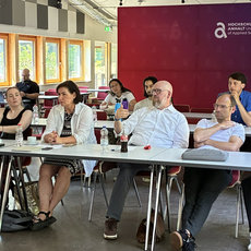 Gruppe von etwa zwölf Personen sitzt an einem langen Tisch in einem hellen Raum, diskutiert und wirkt konzentriert. Im Hintergrund ein Banner mit Logo und Schriftzug „International Academy“.