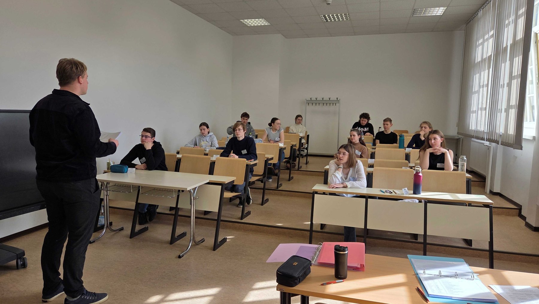 Ein Dozent steht vor einer Gruppe von Schülern im Klassenzimmer und hält eine Präsentation oder ein Blatt Papier. Die Schüler sitzen an Tischen mit Stühlen, einige haben Notizblöcke oder Stifte vor sich.