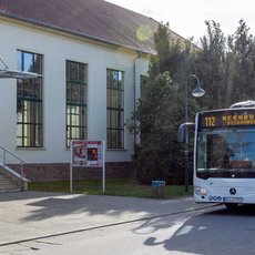 A white bus with the number 112 is parked in front of a light-colored building with large windows. The building has a staircase leading to its entrance and a sign indicating it might be a public facility. The bus displays a route to "Merzbusch." The scene appears to be in a quiet, possibly suburban area.