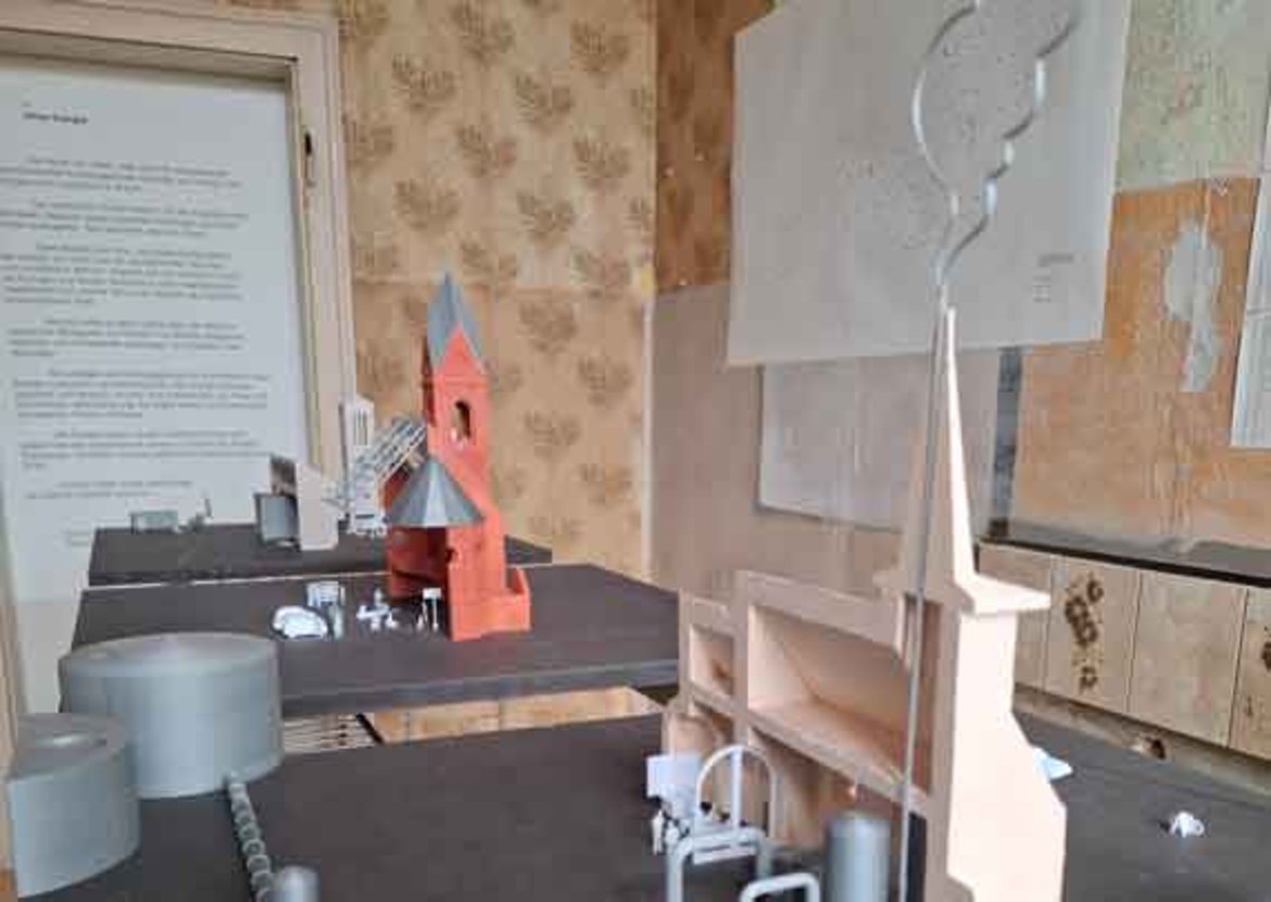 Ausstellungstisch mit Modell eines roten Windradhauses mit grauer Windturbine daneben. Im Hintergrund zwei große, unfertige Skulpturen aus Gips oder Ton, eine davon eine menschliche Figur mit langem Hals.