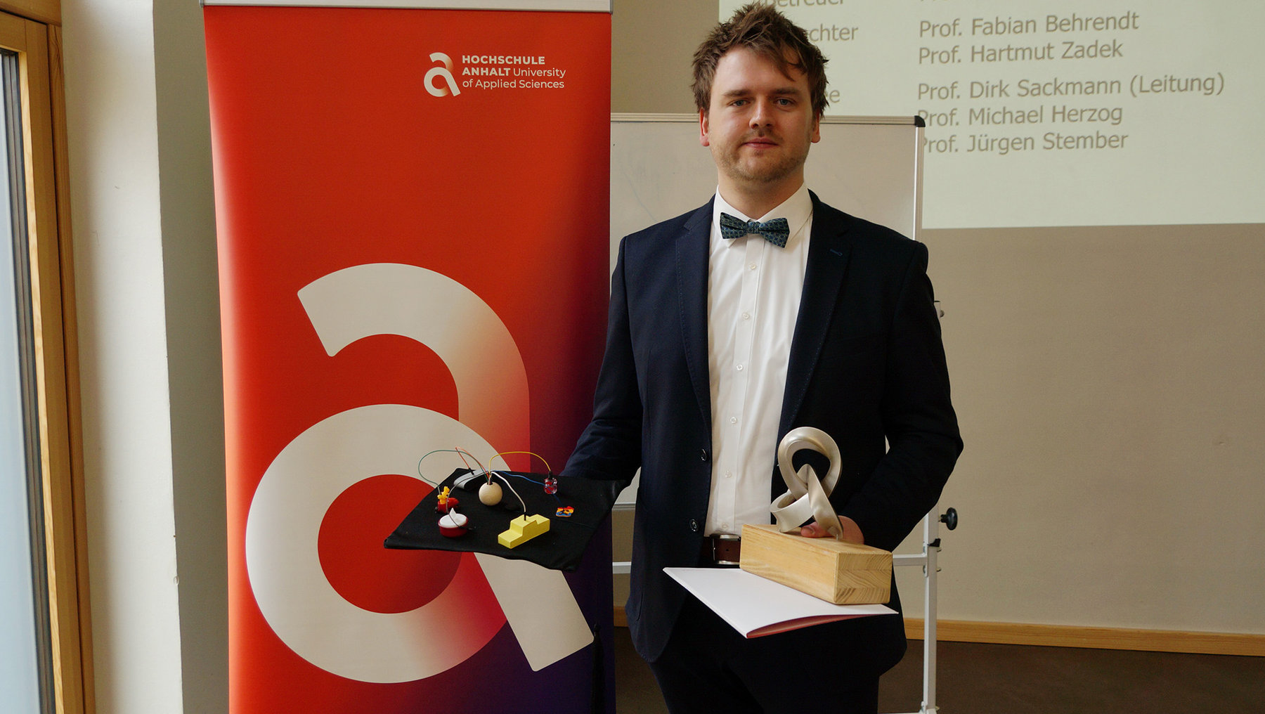 Ein Mann in Anzug mit Weste und Fliege hält ein Modell eines Geräts mit zwei runden Scheiben und einem Kabel, umgeben von einer kleinen Platte mit Obst. Er steht vor einem Banner der Hochschule Aalen.