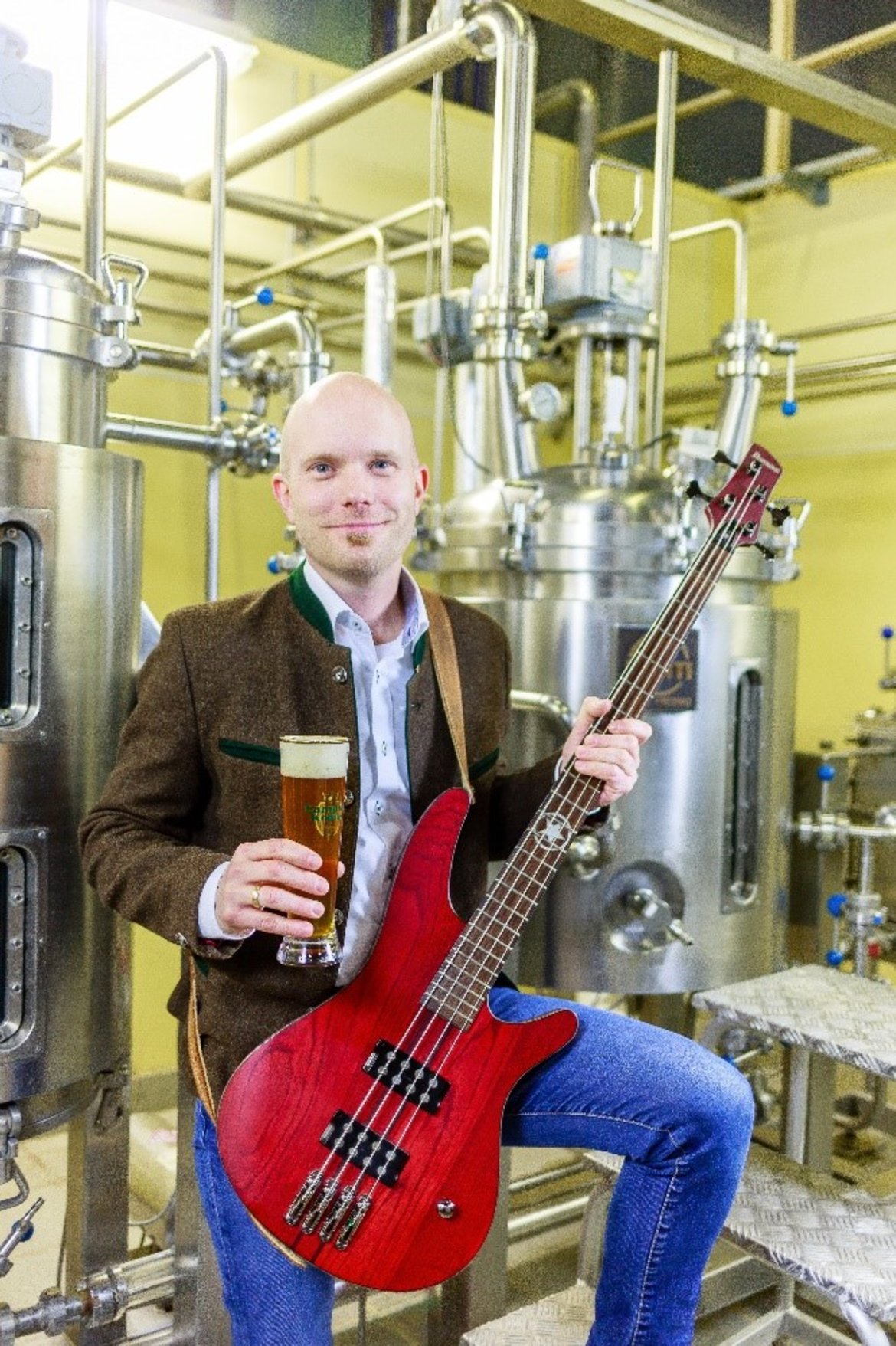 Ein Mann hält eine Bassgitarre in einer Brauerei und trinkt ein Bier. Er trägt einen braunen Blazer, ein blaues Hemd und eine grüne Krawatte. Im Hintergrund sind Braugefäße und Rohrleitungen zu sehen.