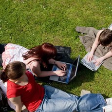 Three people are lying on the grass outdoors, each using a laptop. They are relaxed, with casual clothing and personal items like bags and shoes visible. The scene suggests a study or work session in a park or open area on a sunny day.