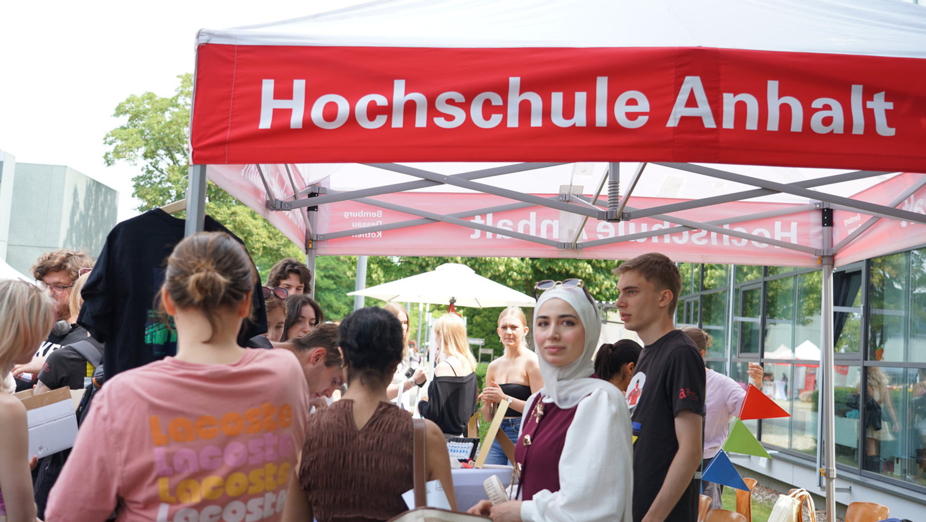 Ein Informationsstand der Hochschule Anhalt mit mehreren Personen, die sich unter einem roten Zelt mit weißer Beschriftung aufhalten. Eine Person im Vordergrund hält Flyer oder Broschüren und spricht mit anderen.