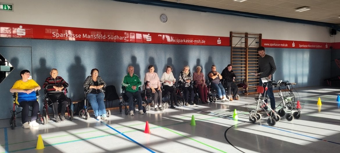 Gruppe von Menschen mit Rollstühlen und einem Mann mit Fahrrad in einer Sporthalle. Der Mann steht vor ihnen und hält ein Fahrrad. Im Hintergrund sind Markierungen auf dem Boden zu sehen, die eine Übungsstrecke andeuten.