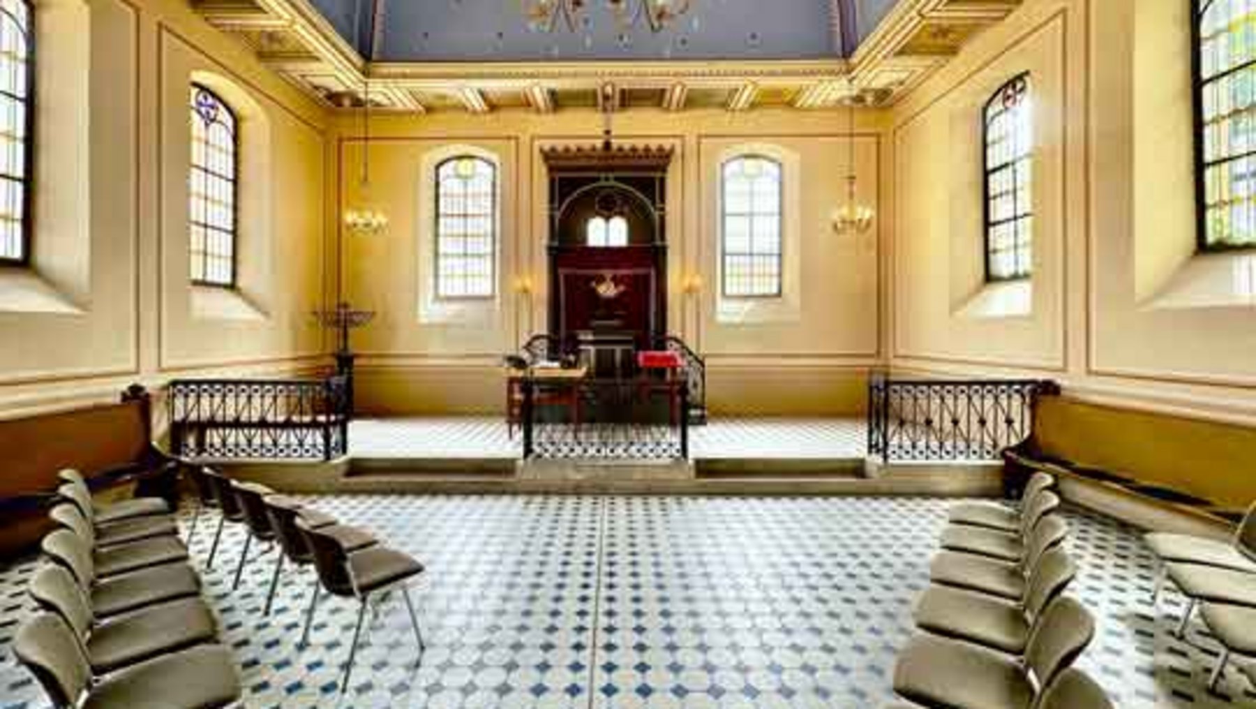 Raum mit hellgelben Wänden und blau-weißem Bodenmuster, typisch für eine Synagoge. Im Zentrum ein Podest mit Lesetisch und Torah-Arkade, flankiert von zwei Treppenaufgängen. An den Wänden große Fenster, an der Decke ein Kronleuchter. Mehrere Reihen mit grauen Sitzbänken stehen symmetrisch angeordnet.
