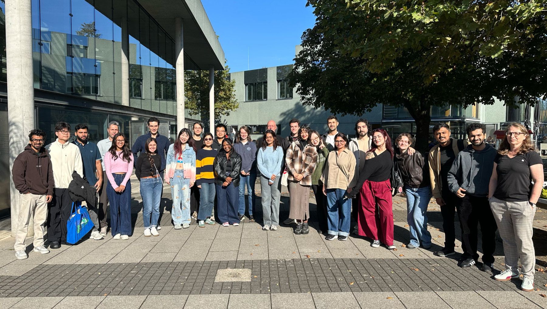 Gruppe von etwa 20 Personen steht lächelnd vor einem modernen Gebäude, vermutlich Studierende oder Teilnehmer einer Veranstaltung. Sie tragen lockere Kleidung, einige halten Taschen oder Jacken. Die Atmosphäre wirkt freundlich und gesellig.