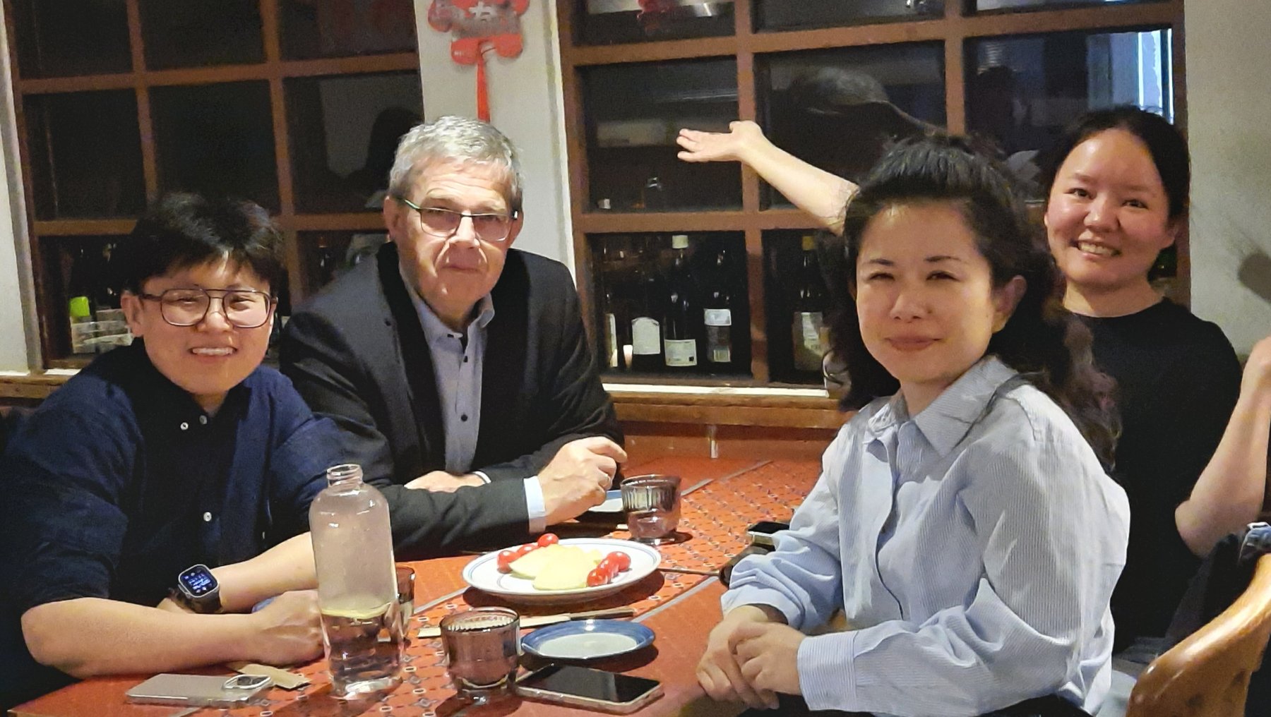 Vier Personen sitzen lächelnd an einem Tisch in einem Restaurant, umgeben von Tellern, Gläsern und Essensresten, während im Hintergrund Regale mit Flaschen zu sehen sind.