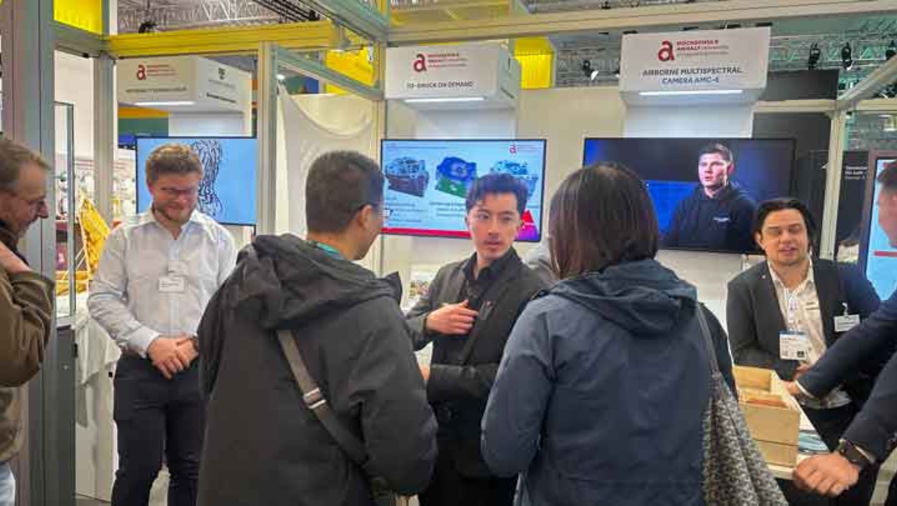 Eine Gruppe von sechs Personen steht in einem Messestand und unterhält sich. Im Hintergrund sind Bildschirme mit Präsentationen und ein Logo mit dem Schriftzug „Airbus Defence and Space“ zu sehen.