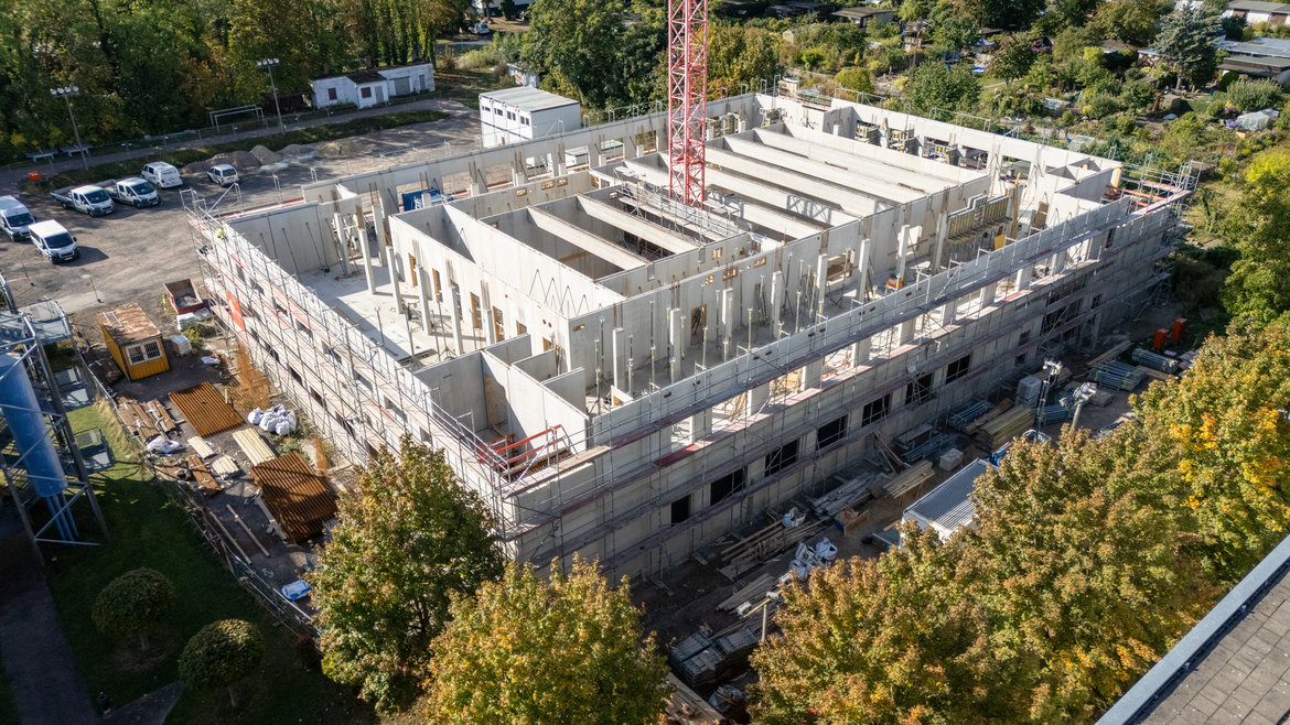 Luftbild eines mehrstöckigen Rohbaus mit Betonwänden und Stahlgerüst, umgeben von Baustelleneinrichtung wie Gerüsten, Containern und Baumaschinen auf einer Baustelle mit angrenzendem Grünbereich.