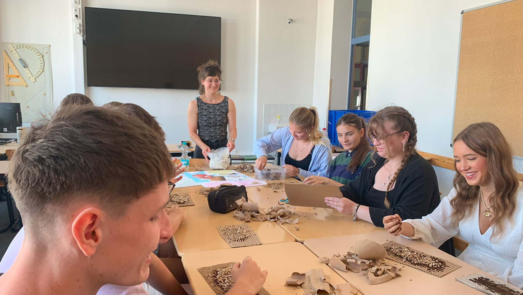 Eine Gruppe von Schülern und einer Hochschulmitarbeiterin sitzt um einen Tisch und untersucht Pilz-Myzel. Alle sind konzentriert und arbeiten gemeinsam an einer Aufgabe.