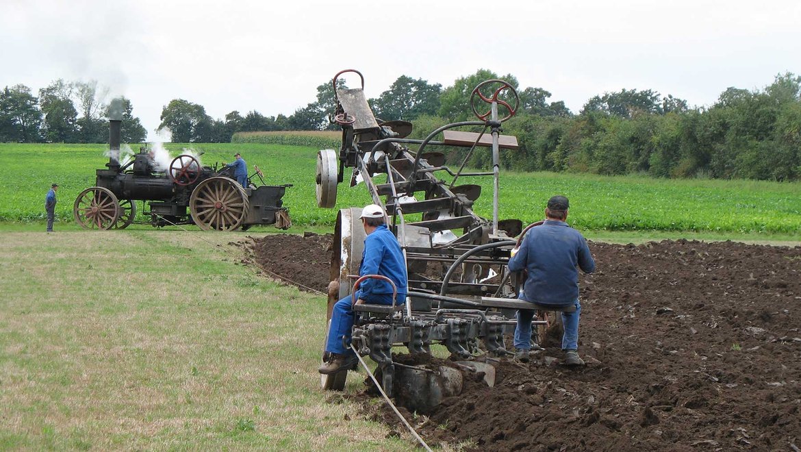 Zwei Männer bedienen eine historische Dampfpflugmaschine auf einem Feld, während im Hintergrund ein Dampftraktor mit dampfender Abgaswolke zu sehen ist. Die Maschine bearbeitet den Ackerboden.