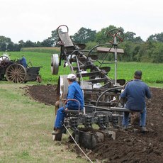 Zwei Männer bedienen eine historische Dampfpflugmaschine auf einem Feld, während im Hintergrund ein Dampftraktor mit dampfender Abgaswolke zu sehen ist. Die Maschine bearbeitet den Ackerboden.