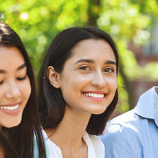Four smiling young people, two women and two men, walking together outdoors in a park, enjoying a sunny day with greenery around them.