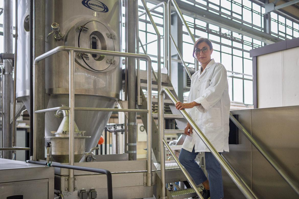 Eine Person in weißem Laborkittel steht lächelnd auf einer Metalltreppe in einer modernen Produktionsanlage mit großen Edelstahltanks und Rohrleitungen, vermutlich in einer Brauerei oder Lebensmittelproduktion.