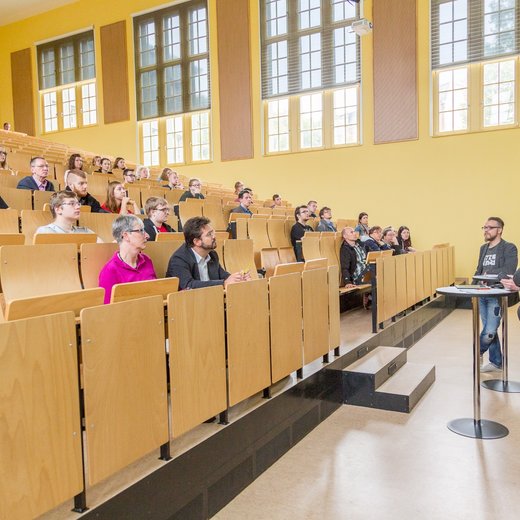 Ein Vortragssaal mit mehreren Reihen von Sitzplätzen, in denen Menschen sitzen und einem Vortragenden zuhören. Der Vortragende steht vorne und spricht an einem Pult. Die Wände sind gelb und es gibt große Fenster, die Licht hereinlassen.