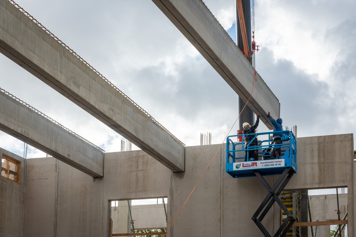 Zwei Bauarbeiter auf einer blauen Hebebühne montieren eine große Betonplatte an einer Rohbaukonstruktion. Die Platte wird mit Seilen positioniert, während einer der Arbeiter sie führt. Im Hintergrund sind Stahlbewehrungen und weitere Bauelemente sichtbar.