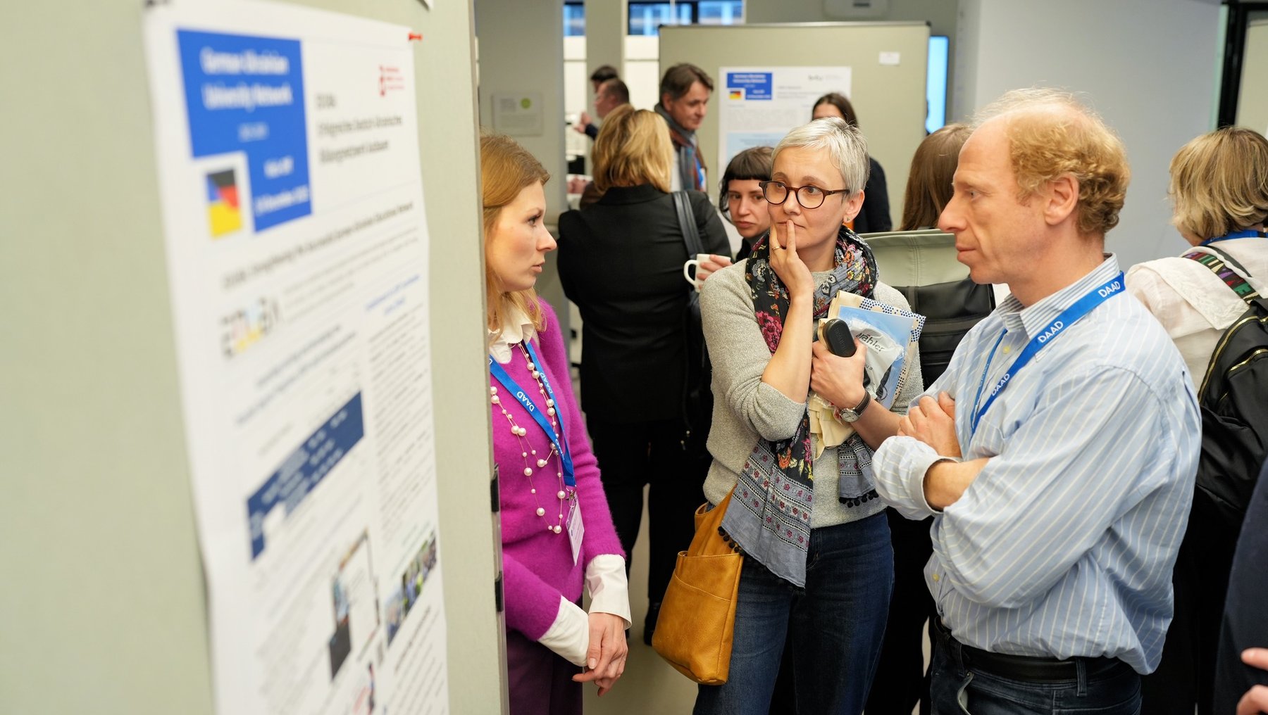 Eine Gruppe von Menschen steht in einem Raum und diskutiert vor einem Poster. Die Personen tragen Namensschilder und scheinen an einer Konferenz oder einem Seminar teilzunehmen. Das Poster enthält Text und Bilder, die wahrscheinlich das Thema der Diskussion darstellen.