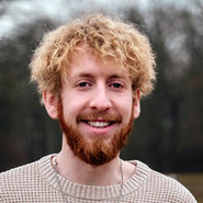 Lächelnder Mann mit kurzem, lockigem blondem Haar und rotem Vollbart, der einen hellbeigen Pullover trägt.