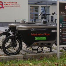 Ein Lastenfahrrad mit dem Aufdruck "Friedheim Filzer" und "Raiffeisen" steht vor einem Schild mit der Aufschrift "Station Campus Köthen". Im Hintergrund ist ein Gebäude mit dem Logo der Hochschule Anhalt zu sehen. Das Schild enthält Informationen zur Ausleihe des Lastenfahrrads.