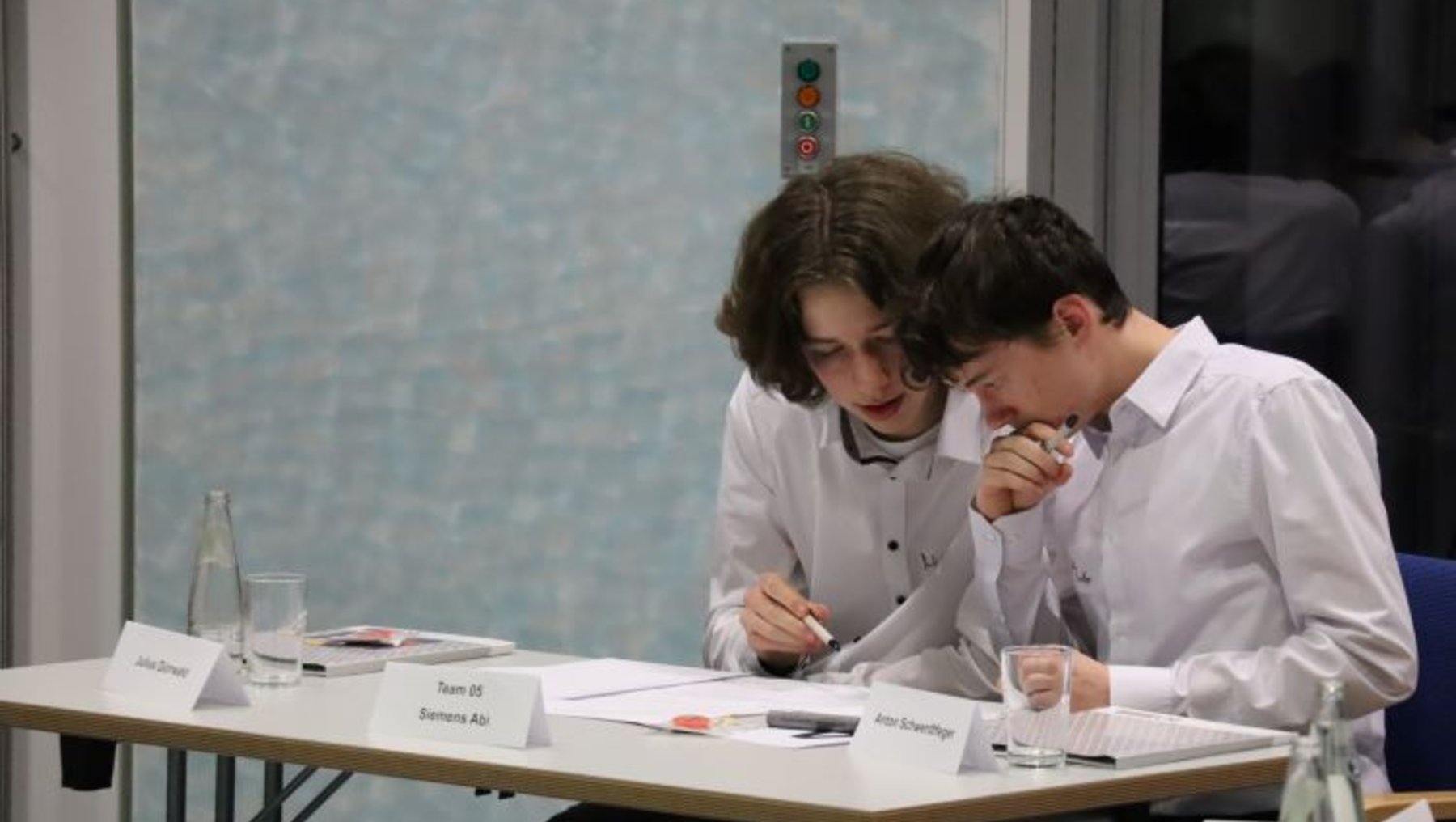 Two individuals in white shirts are seated at a table, engaged in a discussion or working together. They appear to be reviewing documents or taking notes. Nameplates on the table indicate their names and team affiliation. The setting seems to be a formal meeting or collaborative work environment.