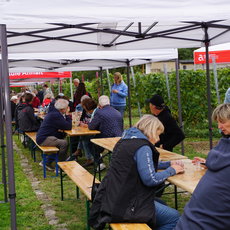 Draußen auf einem Markt oder Fest sitzen mehrere Menschen an langen Holzbänken unter einem großen Zelt. Sie unterhalten sich, essen oder schreiben etwas auf. Im Hintergrund stehen weitere Besucher und ein Infostand mit dem Schriftzug „Hochschule Anhal“.