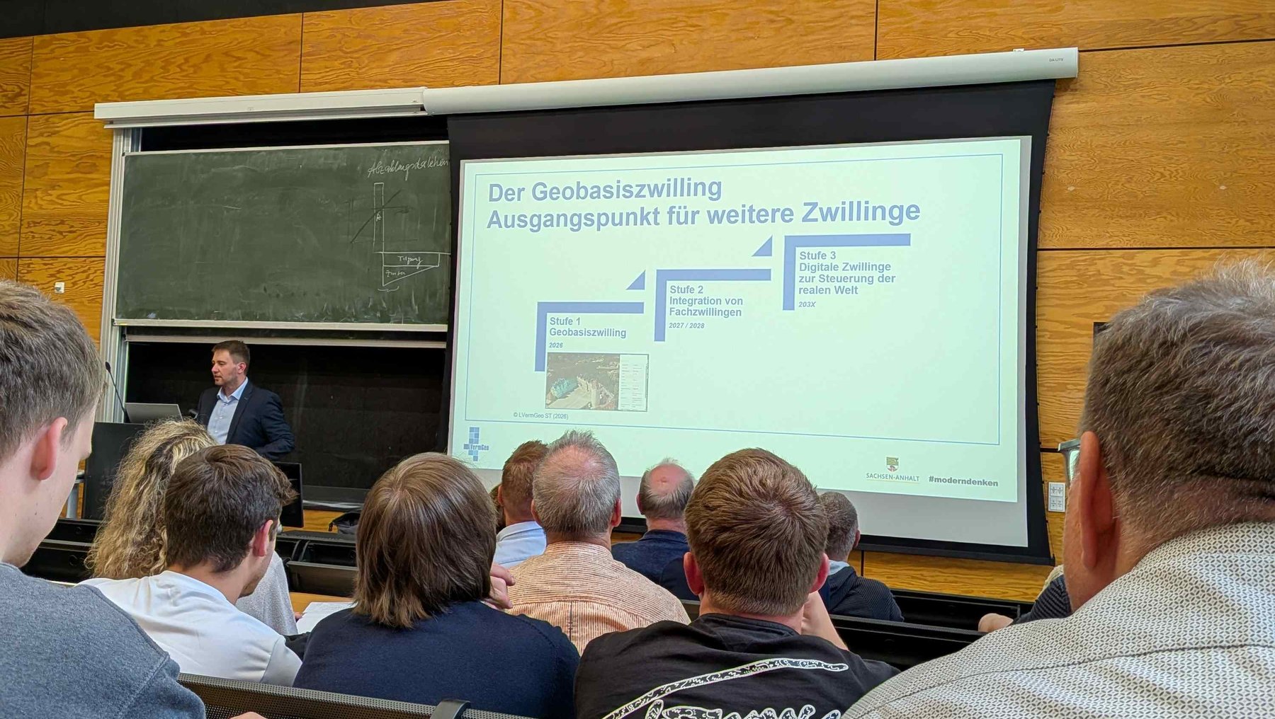 Ein Vortragsraum mit mehreren Zuhörern, die einem Redner auf einer Bühne zuhören. Auf dem Bildschirm wird eine Präsentation mit dem Titel „Der Geobasiswilling Ausgangspunkt für weitere Zwillinge“ angezeigt, die sich mit digitalen Zwillingen und Geodaten befasst.