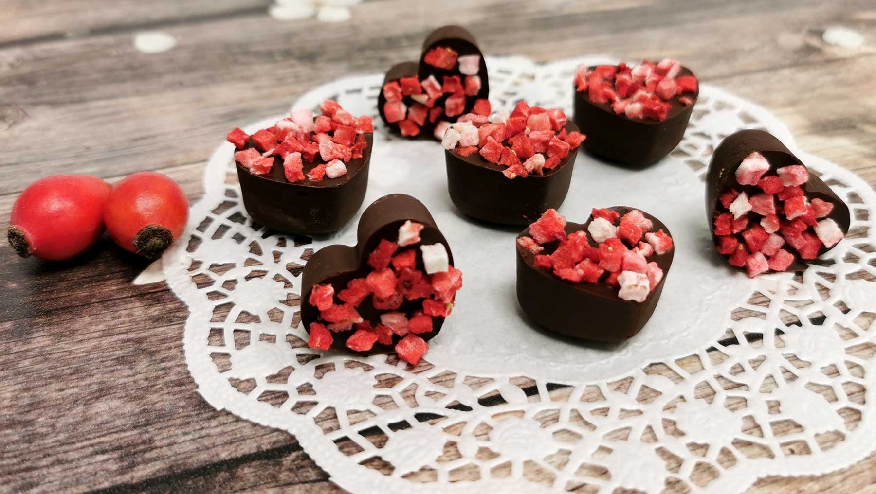 Sechs herzförmige Schokoladenhüllen mit roter Füllung aus gehackten Früchten und weißen Stückchen, arrangiert auf einem Spitzenuntersetzer. Daneben liegen zwei rote Hagebutten.