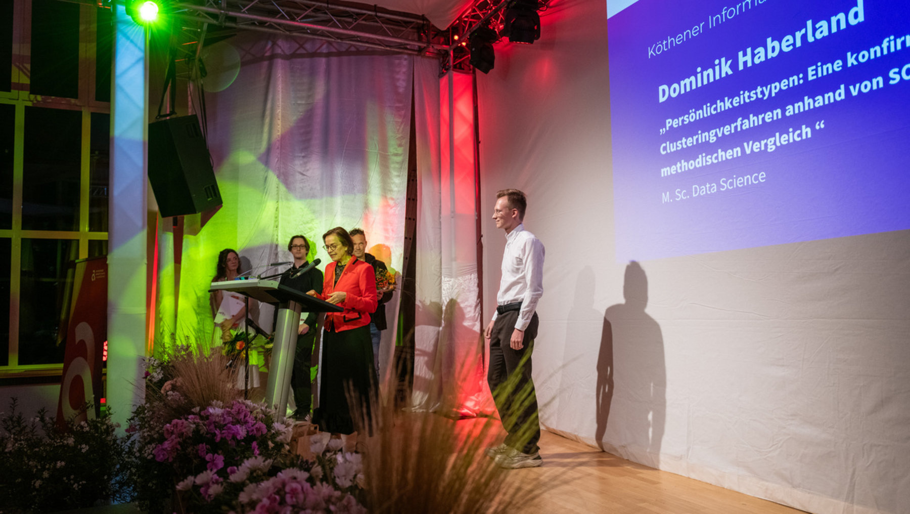 Auf einer Bühne erhält Dominik Haberland für seine wissenschaftliche Arbeit zum Thema Persönlichkeitstypen und Clusteringverfahren einen Preis. Er steht rechts, während links mehrere Personen an einem Podium mit Blumenarrangement sprechen. Im Hintergrund ist eine Projektion mit seinem Namen und dem Titel seiner Masterarbeit zu sehen.