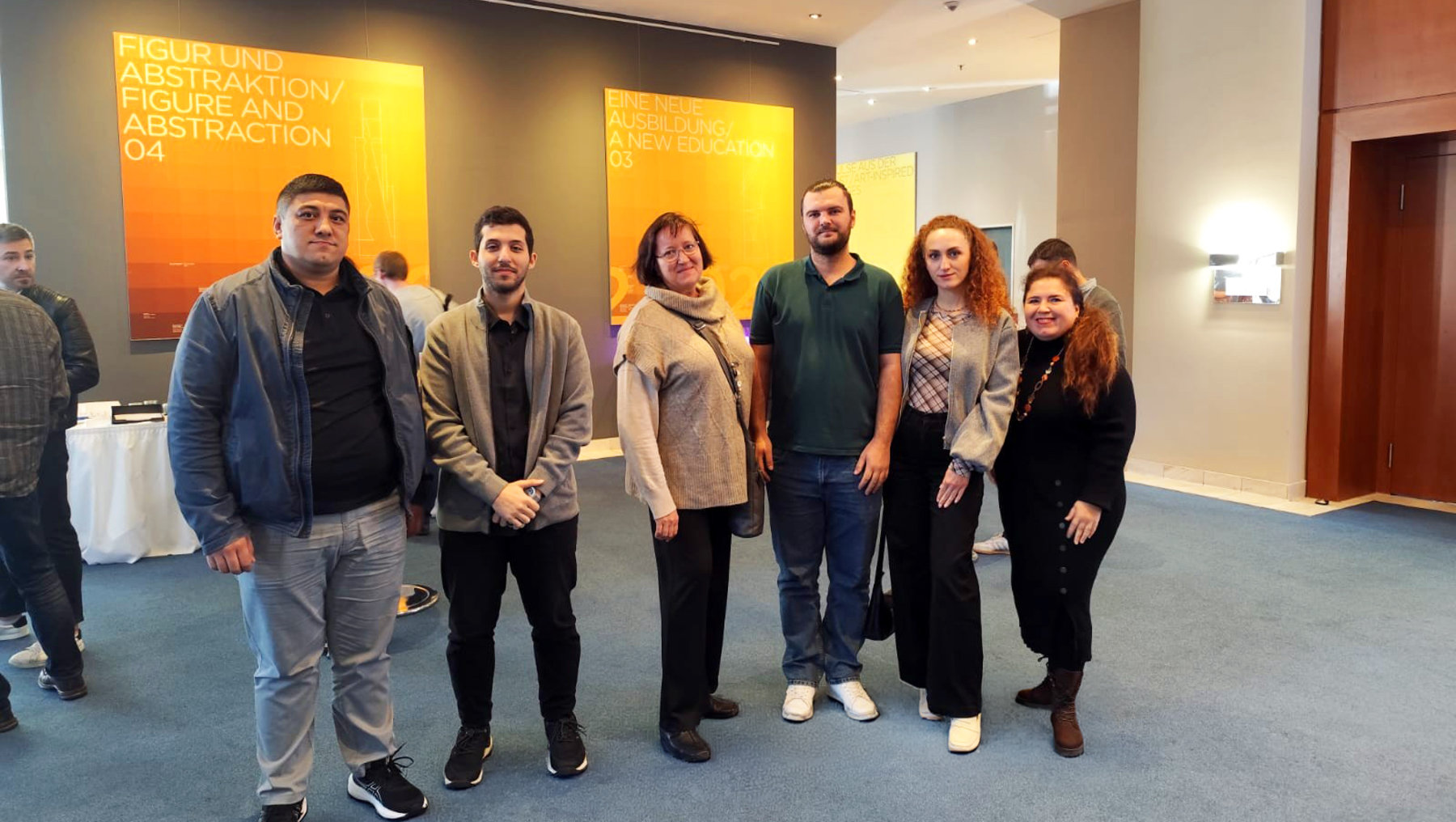 Fünf Personen stehen lächelnd in einer Ausstellungshalle und posieren für ein Gruppenfoto. Im Hintergrund sind gelbe Wände mit dem Titel einer Ausstellung zu abstrakter Kunst zu sehen.