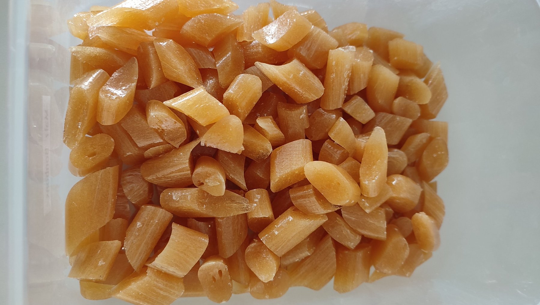Gewürfelte, glasierte Ingwerstücke in einem durchsichtigen Behälter. Die Stücke sind goldbraun und glänzend, was auf eine Zuckerglasur oder ähnliche Zubereitung hindeutet.
