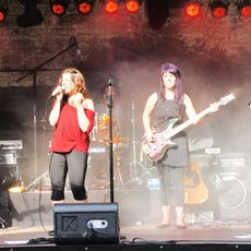 A band performs on stage with four members: two guitarists, a singer, and a bassist. The singer stands at the center with a microphone, while the other three play their instruments. The stage is lit with red lights, creating a vibrant atmosphere.