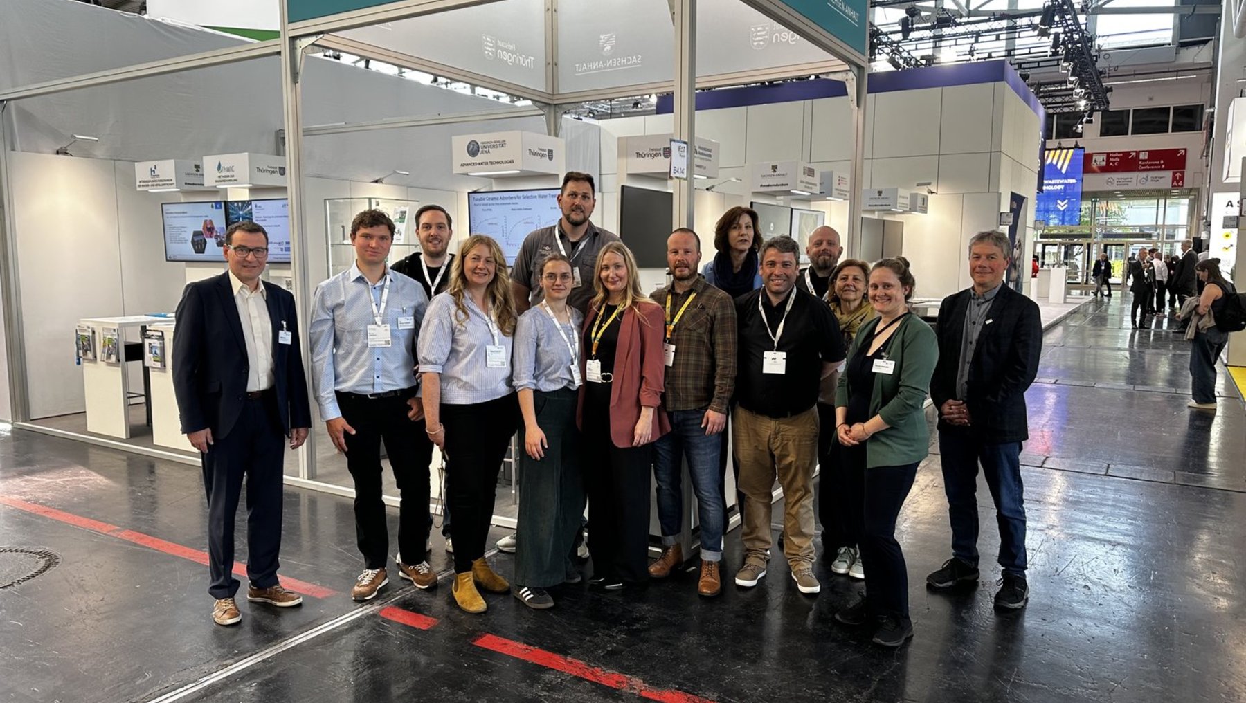 Gruppe von 14 Personen, die auf einer Messe vor einem Gemeinschaftsstand mit Firmenlogos wie Metzen, Thörlberger und Sachsen-Anhalt lächeln und für ein Gruppenfoto posieren.