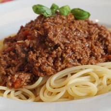A plate of spaghetti topped with a generous serving of meat sauce. Fresh basil leaves are garnished on top, adding a touch of green to the dish. The pasta appears well-cooked and the sauce is rich and hearty.