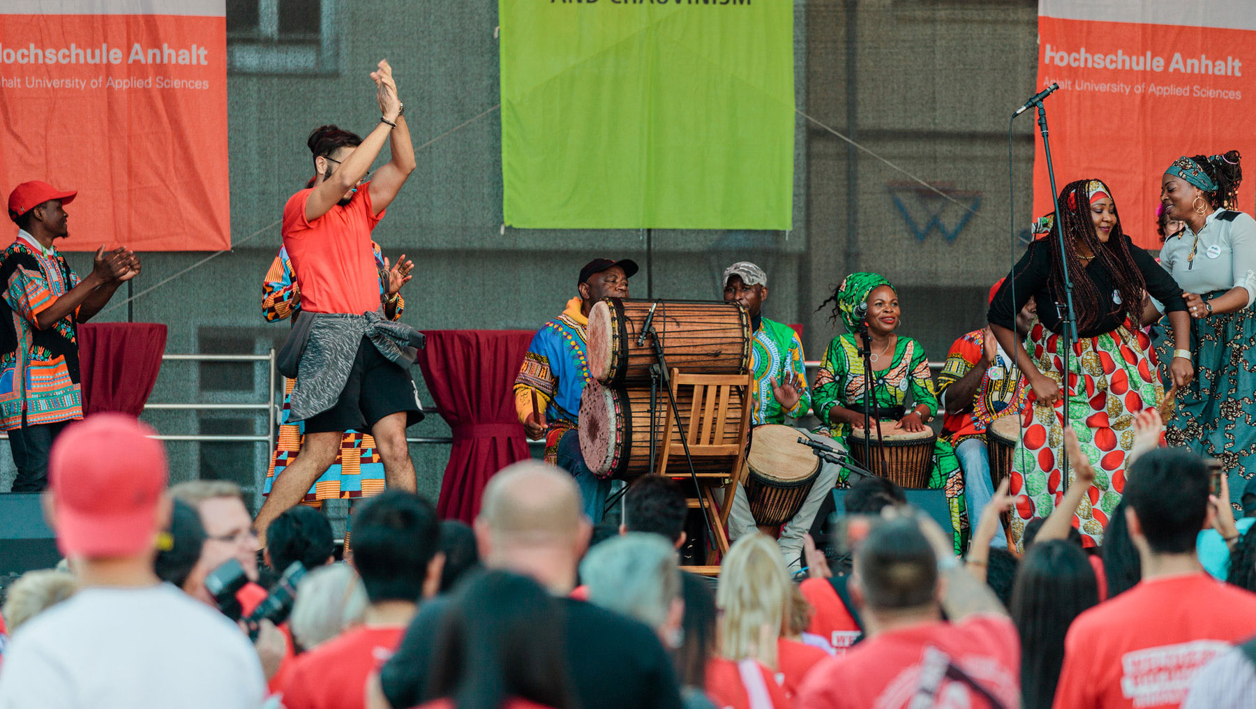 Auf einer Bühne spielt eine Gruppe Menschen traditionelle afrikanische Musik mit Trommeln und Perkussionsinstrumenten. Ein Tänzer in der Mitte feiert mit erhobenen Armen. Das Publikum klatscht begeistert.