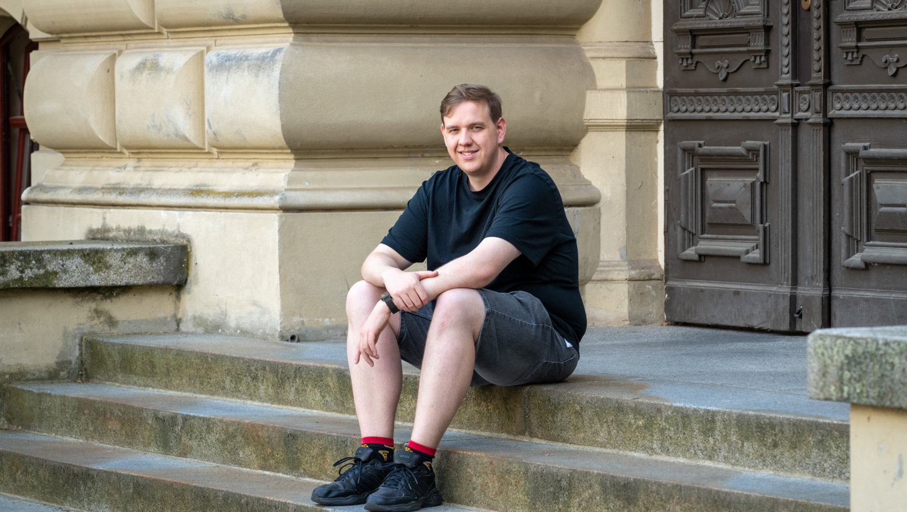 Ein junger Mann sitzt lächelnd auf den Stufen eines historischen Gebäudes mit verzierten Säulen und einer schwarzen Tür, trägt ein schwarzes T-Shirt, kurze graue Hose, rote Socken und schwarze Turnschuhe.