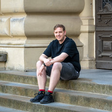 Ein junger Mann sitzt lächelnd auf den Stufen eines historischen Gebäudes mit verzierten Säulen und einer schwarzen Tür, trägt ein schwarzes T-Shirt, kurze graue Hose, rote Socken und schwarze Turnschuhe.