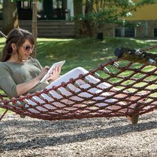 A woman is relaxing in a hammock outdoors, reading a book. She is wearing sunglasses and casual clothing. The hammock is suspended between a tree and a metal stand. The setting appears to be a peaceful garden or backyard area.