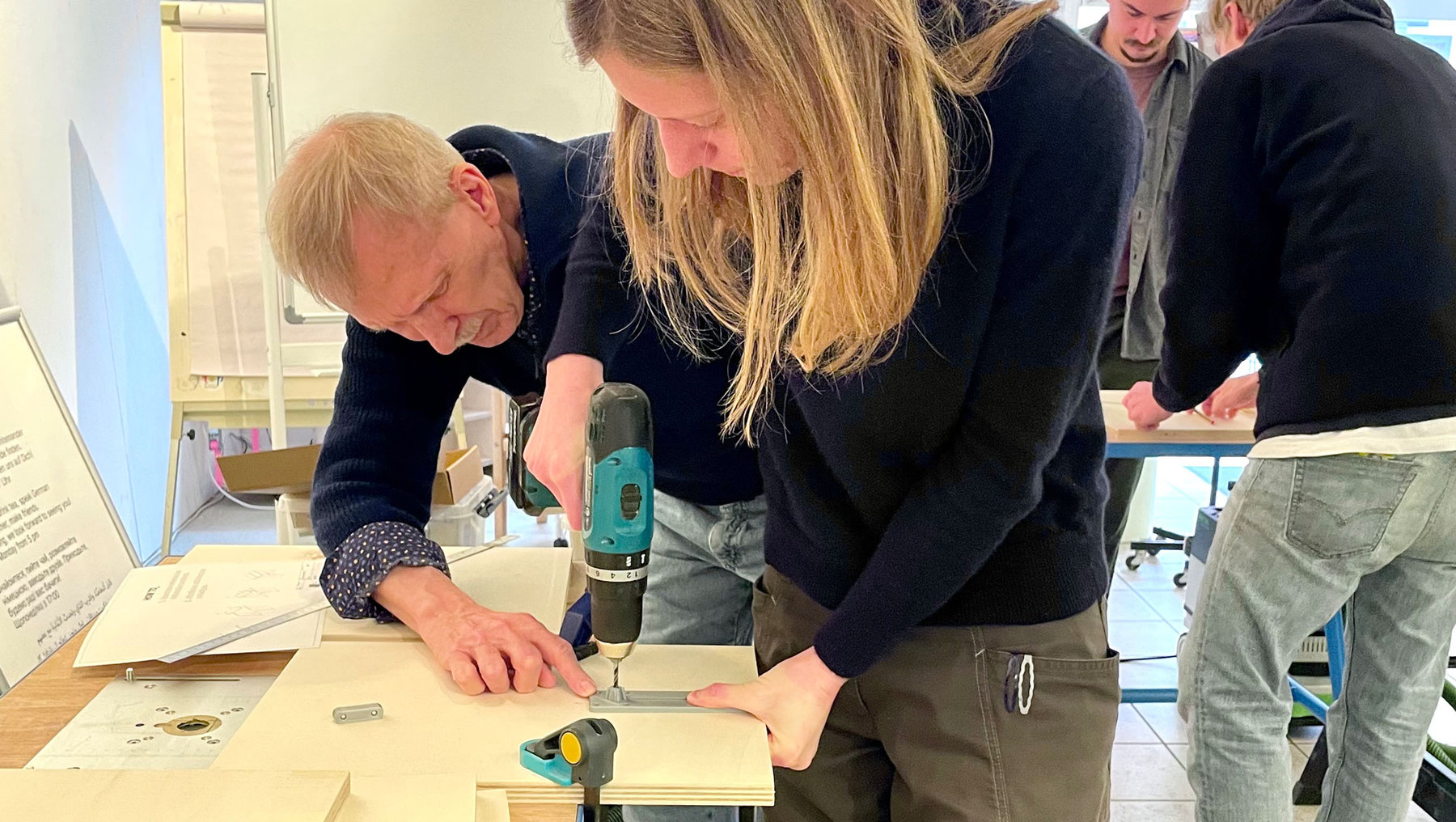 Zwei Personen bohren gemeinsam an einer Werkbank mit einer Bohrmaschine an einem Holzbrett, während andere im Hintergrund zuschauen. Ein Workshop oder praktische Anleitung findet statt.