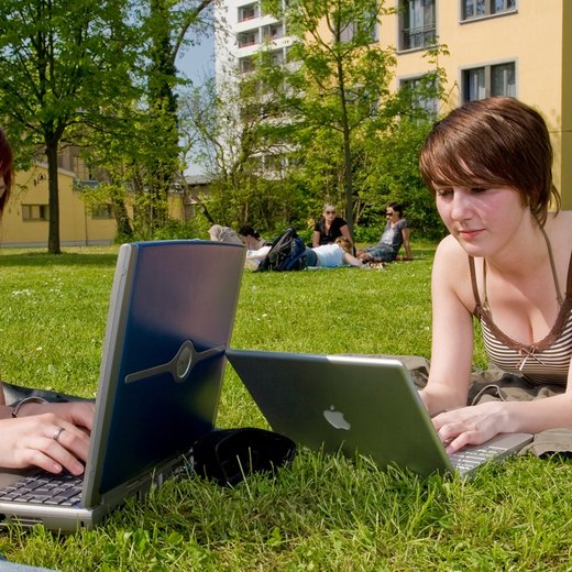 Two women are lying on the grass outdoors, each using a laptop. They appear to be engaged in conversation while working. The background shows a park with trees and a building. The scene suggests a relaxed, collaborative study or work session in a pleasant environment.