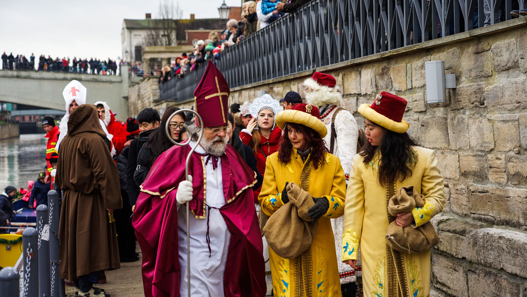 Eine Gruppe von Menschen in historischen und festlichen Kostümen nimmt an einer Parade teil. Die Kostüme umfassen prächtige Roben, Hüte und Accessoires. Die Teilnehmer scheinen in einer festlichen Atmosphäre zu sein, möglicherweise bei einem traditionellen Fest oder einer Feier.