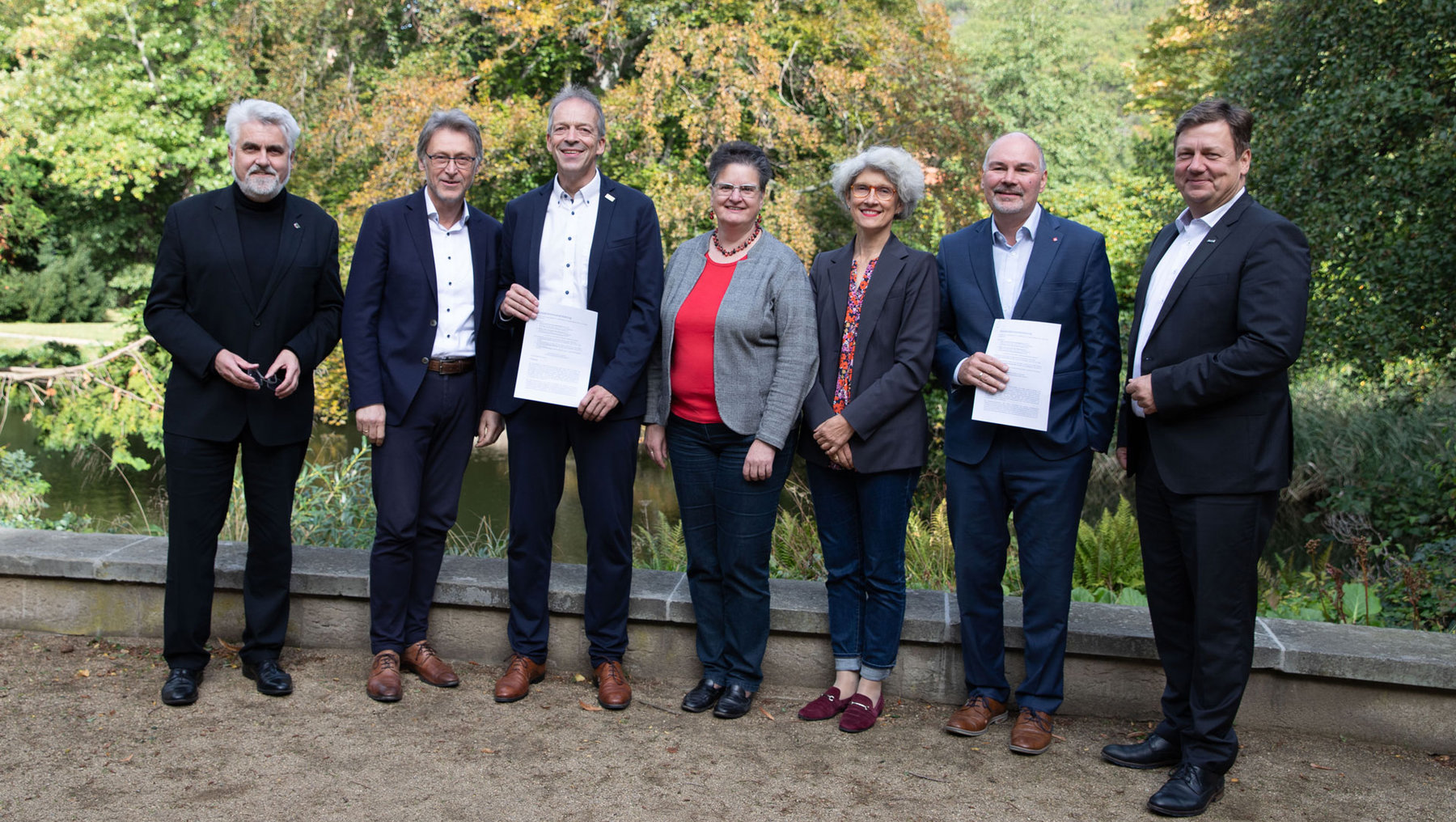 Sechs Personen stehen im Freien und halten jeweils ein Dokument oder eine Urkunde. Sie lächeln und posieren für ein Gruppenfoto, vermutlich bei einer offiziellen Veranstaltung oder Preisverleihung.